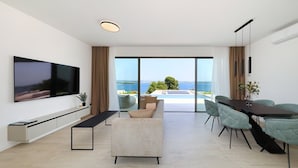 A modern living room with a flat-screen TV, a sofa, a dining table with chairs, and a large sliding glass door leading to a pool and ocean view. - Villa D9 with private pool 1 km from sea (Tkon)