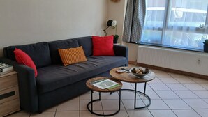 A living room with a sofa, a coffee table, a side table with books, and a window with curtains.