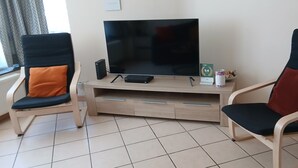 A living room with a TV, two chairs, a small table with a can, and a window with curtains.