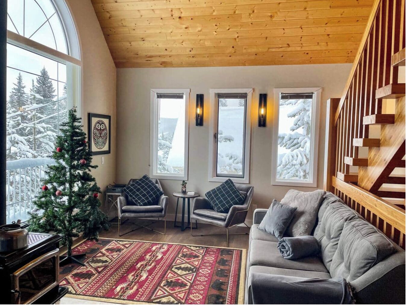 Un salon chaleureux avec une cheminée, un sapin de Noël et une vue sur les arbres enneigés, visible à travers de grandes fenêtres.