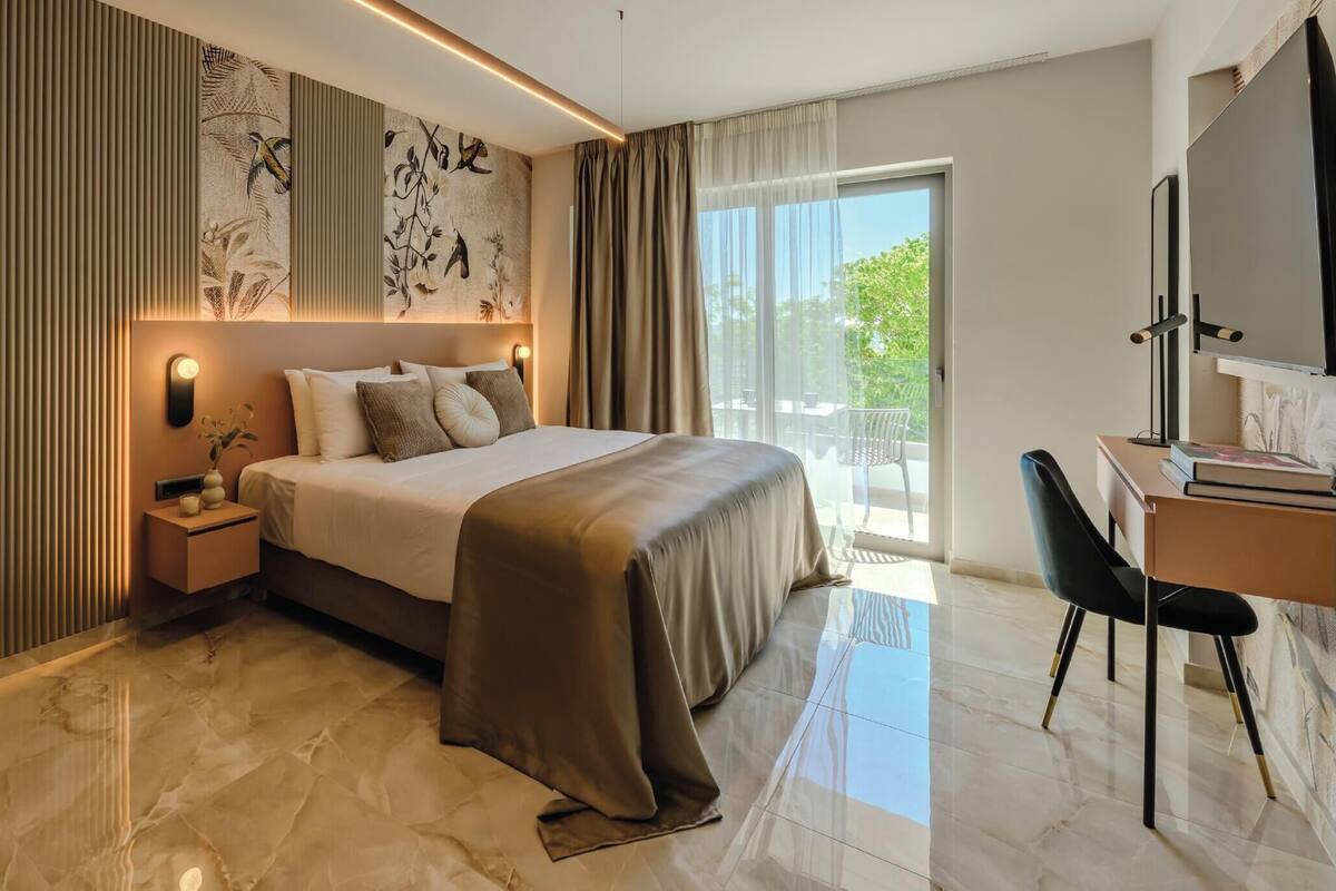 A modern hotel room with a bed, bedside table, lamp, and a desk with a chair.