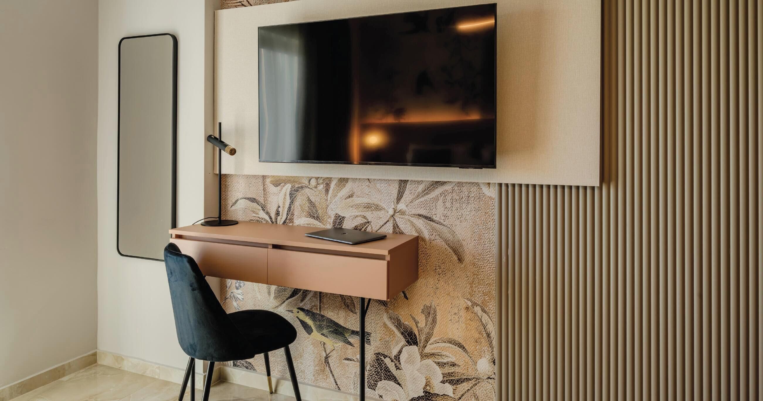A modern room with a flat-screen TV mounted on the wall, a desk with a laptop, a chair, and a large mirror.