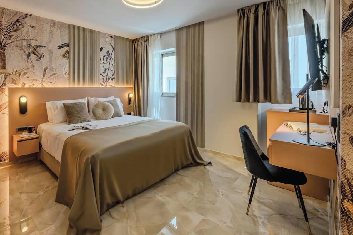 A hotel room with a bed, bedside table, desk, chair, and a large window with curtains.
