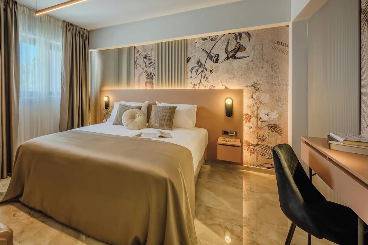 A hotel room with a large bed, a desk, a chair, and a nightstand with a lamp.