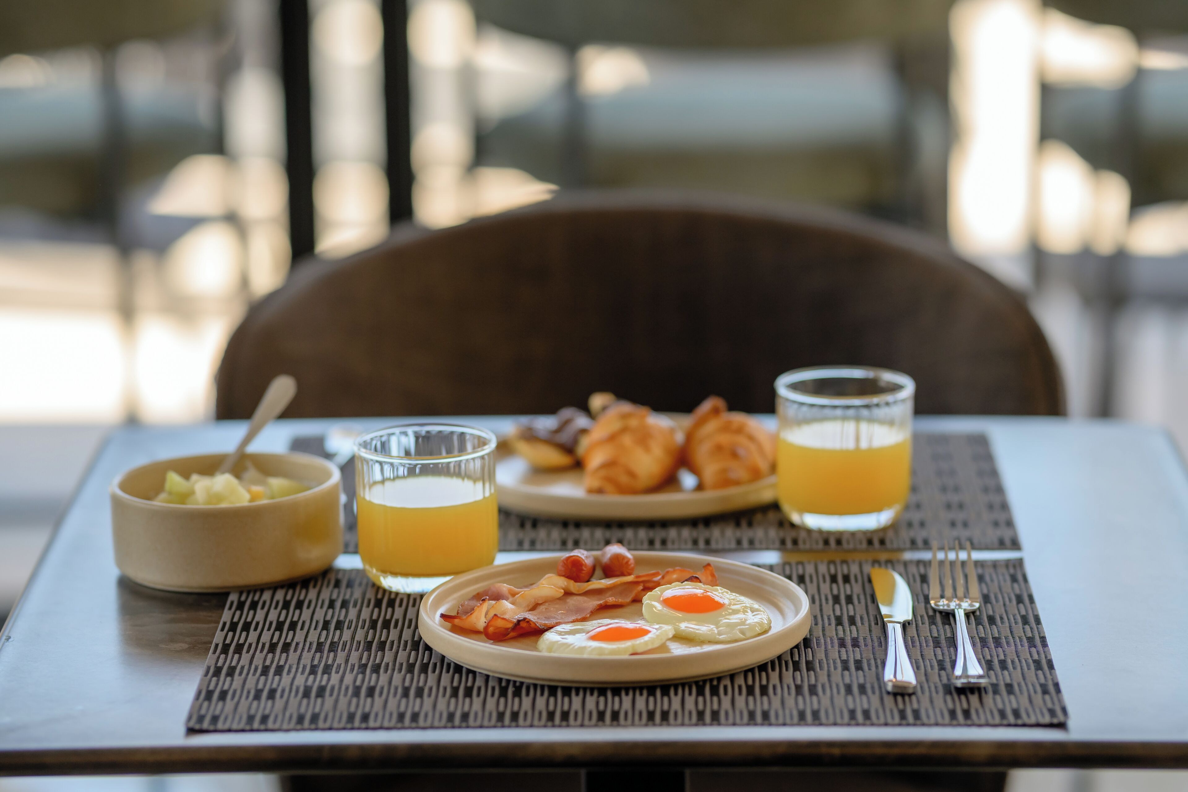 Een ontbijttafel met croissants, eieren, spek, een schaal fruit en twee glaasjes sinaasappelsap.