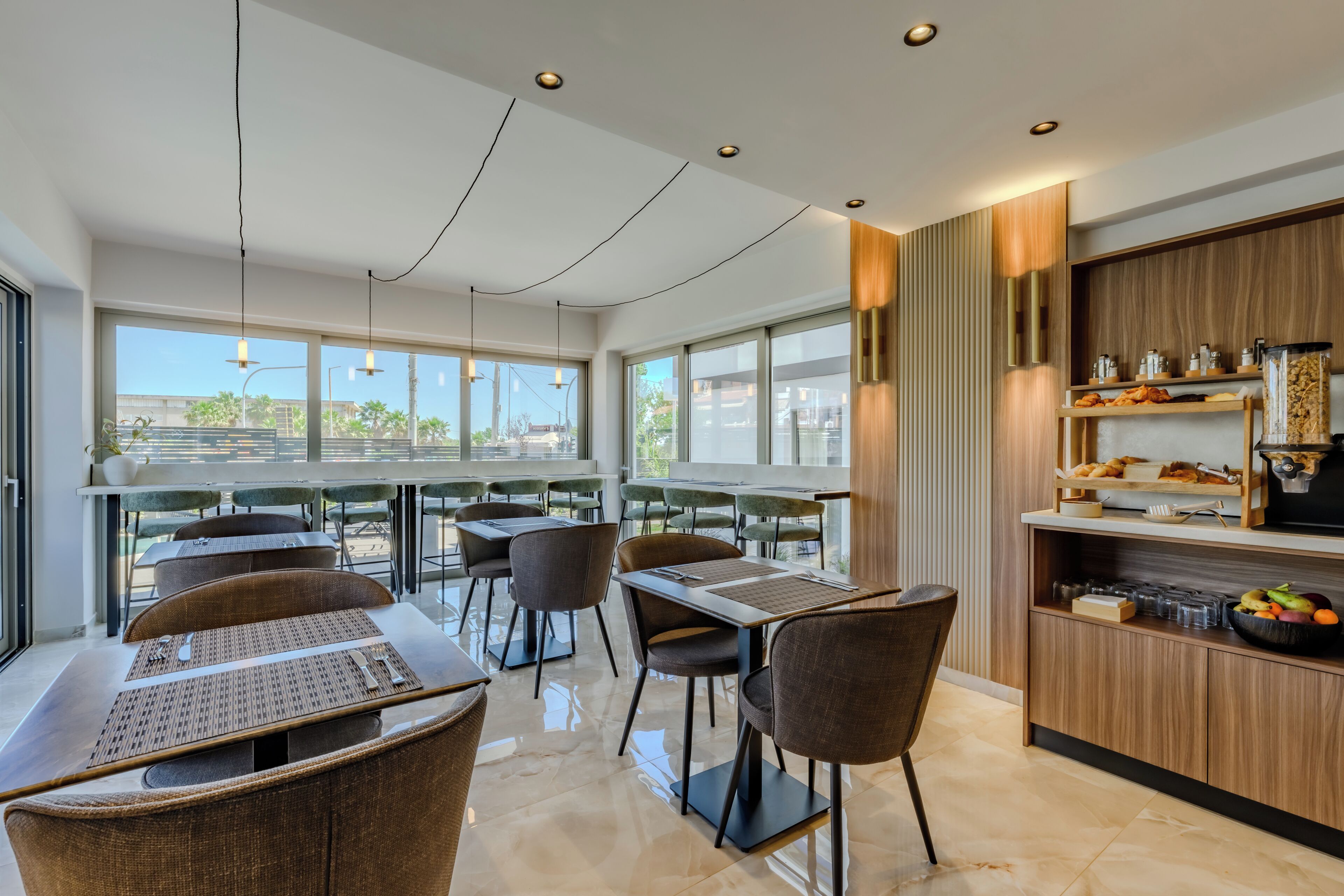 Un moderno comedor con mesas y sillas, una encimera de cocina con alimentos, y ventanas amplias que ofrecen vistas.