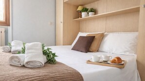 Cama bem arrumada com toalhas dobradas, um tabuleiro com chávenas e um croissant, e uma estante com plantas.