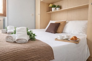 Ein ordentlich bezogenes Bett, zusammengerollte Handtücher, eine Ablage mit Tassen und einem Croissant sowie ein Regal mit Pflanzen.