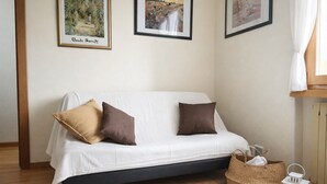 Um sofá branco com duas almofadas castanhas, uma cesta de vime, um candeeiro branco e um livro aberto sobre o soalho de madeira.