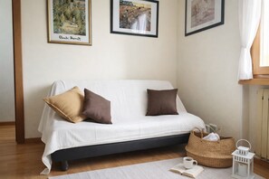 Ein weißes Sofa mit zwei braunen Kissen, ein Korbwannenstuhl, ein weißer Laternen und ein offenes Buch auf Holzboden.