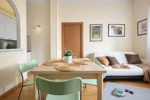 Ein gemütliches Wohnzimmer mit einem Holzesstisch, grünen Stühlen, einer braunen Couch mit Kissen und einer kleinen Topfpflanze.