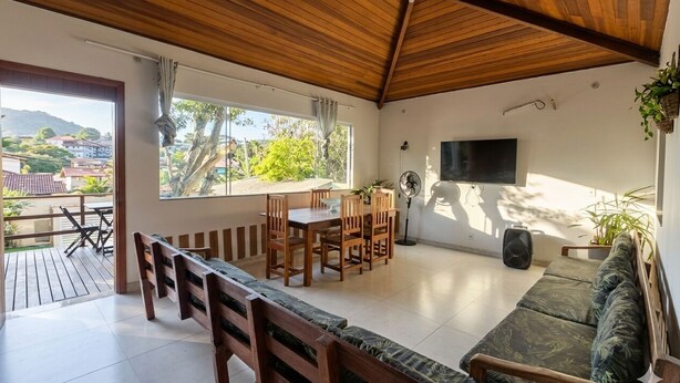 A spacious room with a wooden ceiling, a dining area with a table and chairs, a sofa, a TV mounted on the wall, and a balcony with a view of the surrounding area. - House in Geribá, 5 minutes from the beach, with pool, sleeps 26. (Armação  dos Búzios)