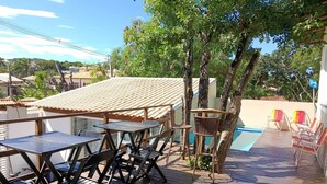 A wooden deck with tables and chairs, a pool, and a view of houses in the background. - House in Geribá, 5 minutes from the beach, with pool, sleeps 26. (Armação  dos Búzios)
