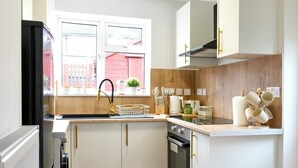 Une cuisine de taille réduite avec des meubles blancs, un plan de travail en bois, un réfrigérateur noir et une fenêtre donnant sur un immeuble rouge.