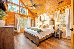 Un dormitorio de cabaña de madera con cama, cortinas, ventilador de techo y ventana con vista a árboles.