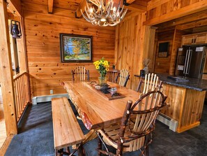 A wooden interior with a dining table, chairs, a chandelier, a painting, and a kitchen area.