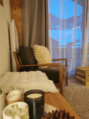 Ein gemütliches Wohnzimmer mit einem Holzstuhl, einem weißen Kissen, einem kleinen Tisch mit Kerzen und Blick auf eine verschneite Landschaft durch ein Fenster.