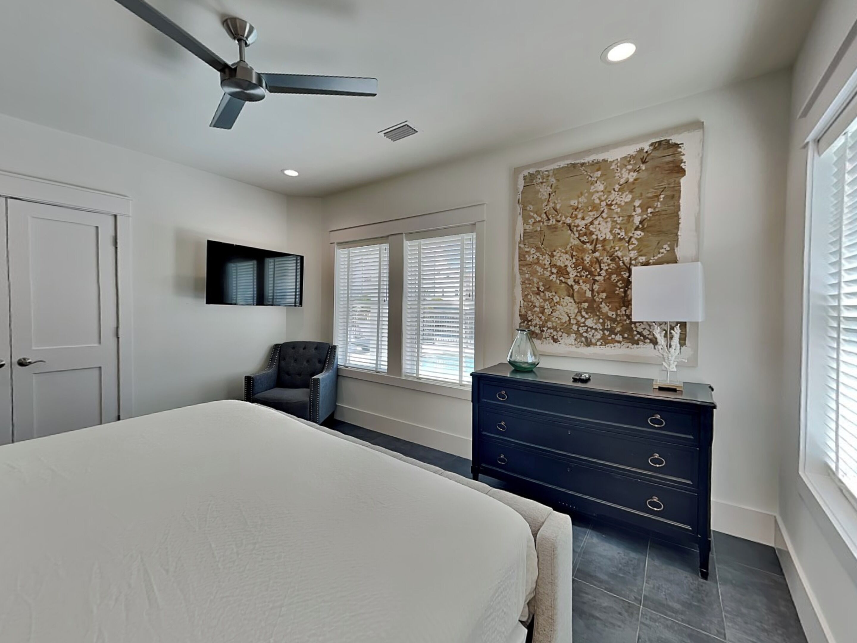A bedroom with a bed, a dresser, a chair, a ceiling fan, and a large window with blinds.