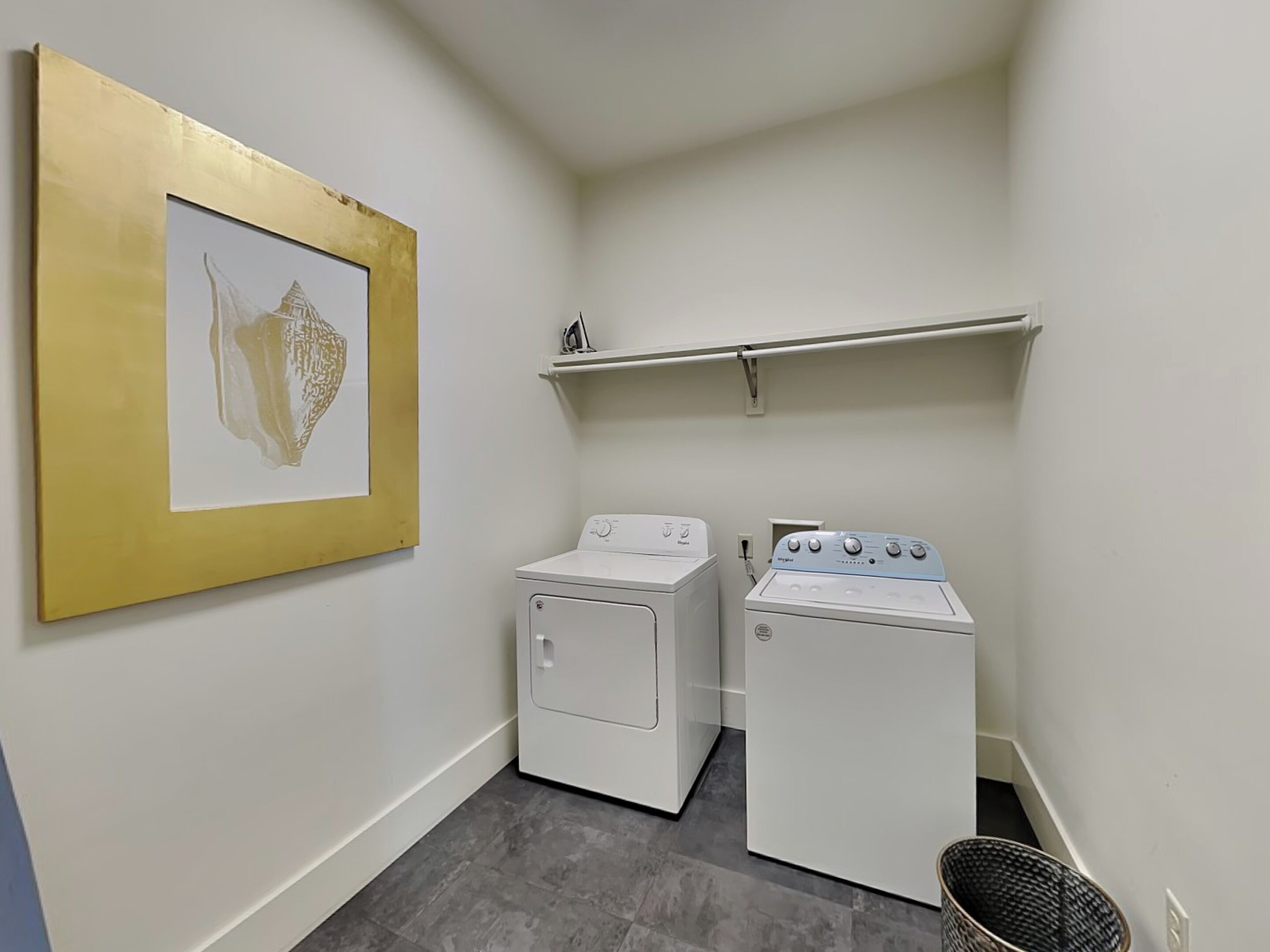 A laundry room with two white washing machines, a shelf above, and a framed picture on the wall.