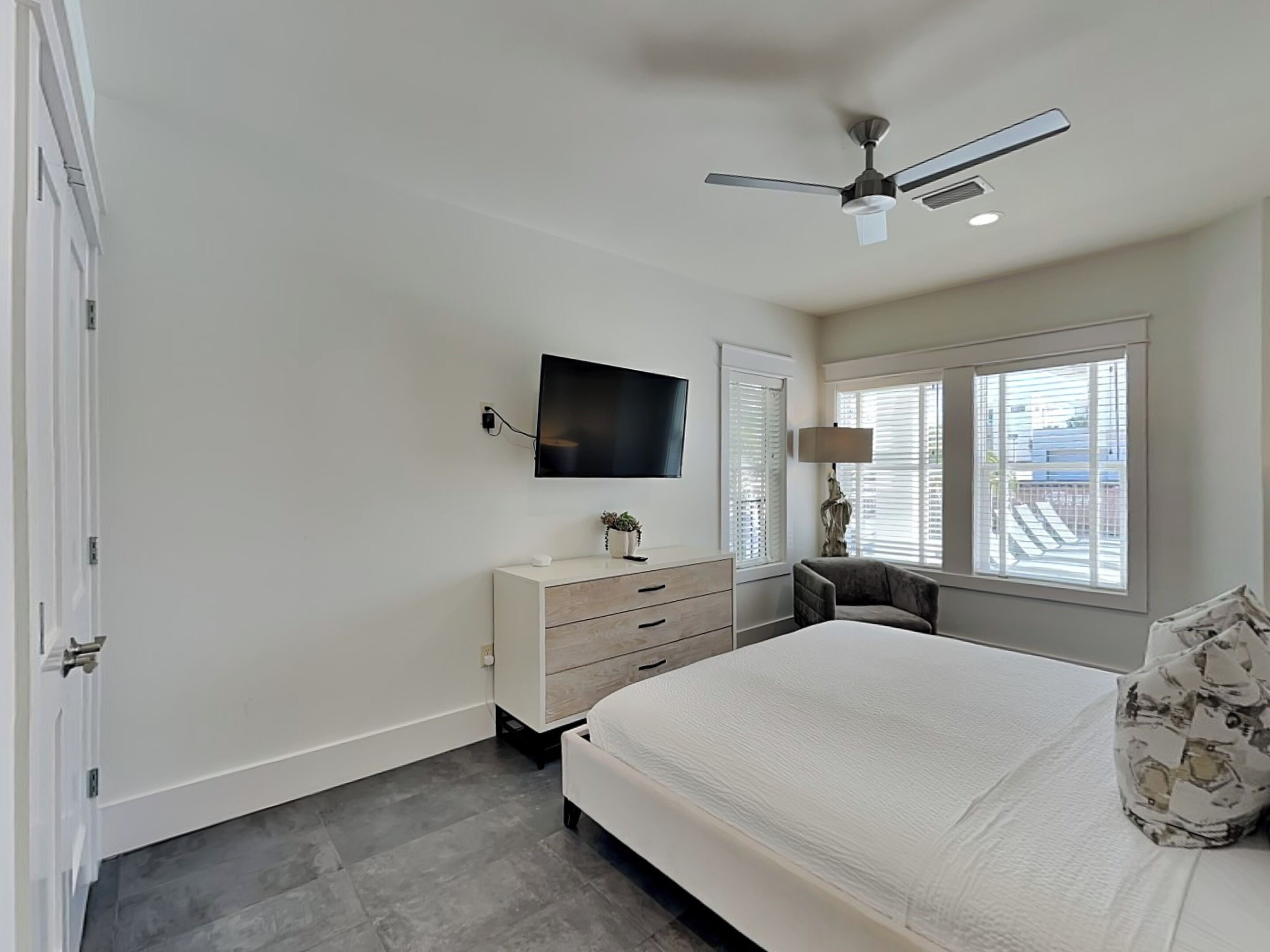 Een slaapkamer met een bed, een dressoir, een televisie, een plafondventilator en een raam met jaloezieën.
