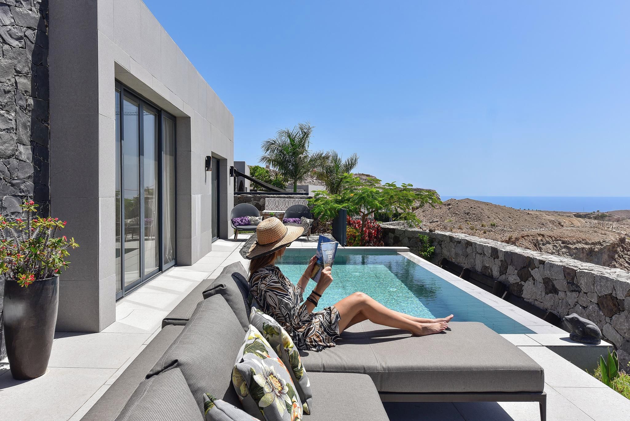 A person relaxing on a grey outdoor sofa by a swimming pool with a view of the sea.
