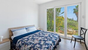 Un dormitorio con cama, una silla, una mesita de noche y un amplio ventanal corredera que da a un espacio exterior.