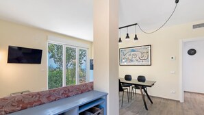 Una habitación moderna con televisión, un banco con cojín floral, zona de comedor con mesa y sillas, y una obra de arte en la pared.