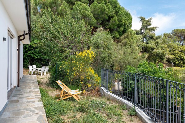 A stone pathway leading to outdoor seating, surrounded by lush greenery and a metal fence.