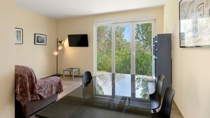 Una habitación moderna con sofá, televisión, mesa de comedor con sillas y un amplio ventanal con vistas a árboles.