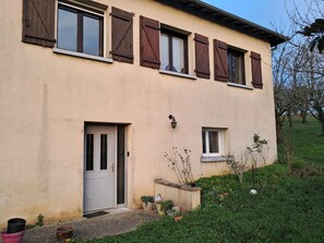 Un bâtiment à deux étages, à la façade beige, avec des volets en bois aux fenêtres, une porte blanche et une petite fenêtre à cadre blanc. On trouve des plantes en pot et un petit jardin devant le bâtiment.