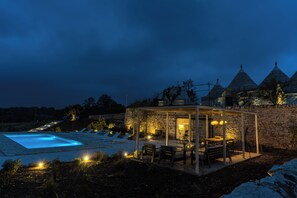 Ein Poolbereich mit Liegestühlen, eine überdachte Sitzgelegenheit mit Tischen und dahinter stehende Steinbauten.