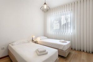 Quarto de hotel com duas camas, luminária de cabeceira, lustre e cortinas leves.