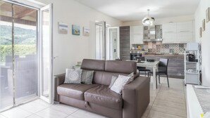 Un salon moderne avec un canapé en cuir marron, une table à manger avec des chaises, un coin cuisine doté de placards et d’un plan de travail, et une porte-fenêtre coulissante donnant sur un balcon.