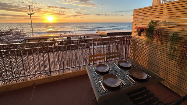 Ein Balkon mit Blick auf den Sonnenuntergang über dem Meer, ein Tisch gedeckt mit Geschirr und Besteck, und eine Holzwand mit hängenden Pflanzen.