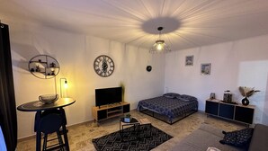 Ein modernes Wohnzimmer mit Bett, Fernseher, Esstisch und einer Deckenleuchte.