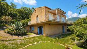 Un edificio a due piani con un balcone, immerso nel verde sotto un cielo sereno.