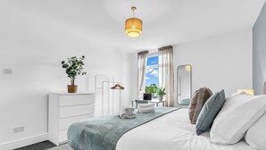 A bedroom with a bed, a dresser, a mirror, a hanging plant, and a window with curtains.