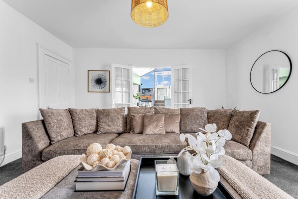 A modern living room with a sectional sofa, a coffee table with decorative items, a large mirror, and a view through an open door.