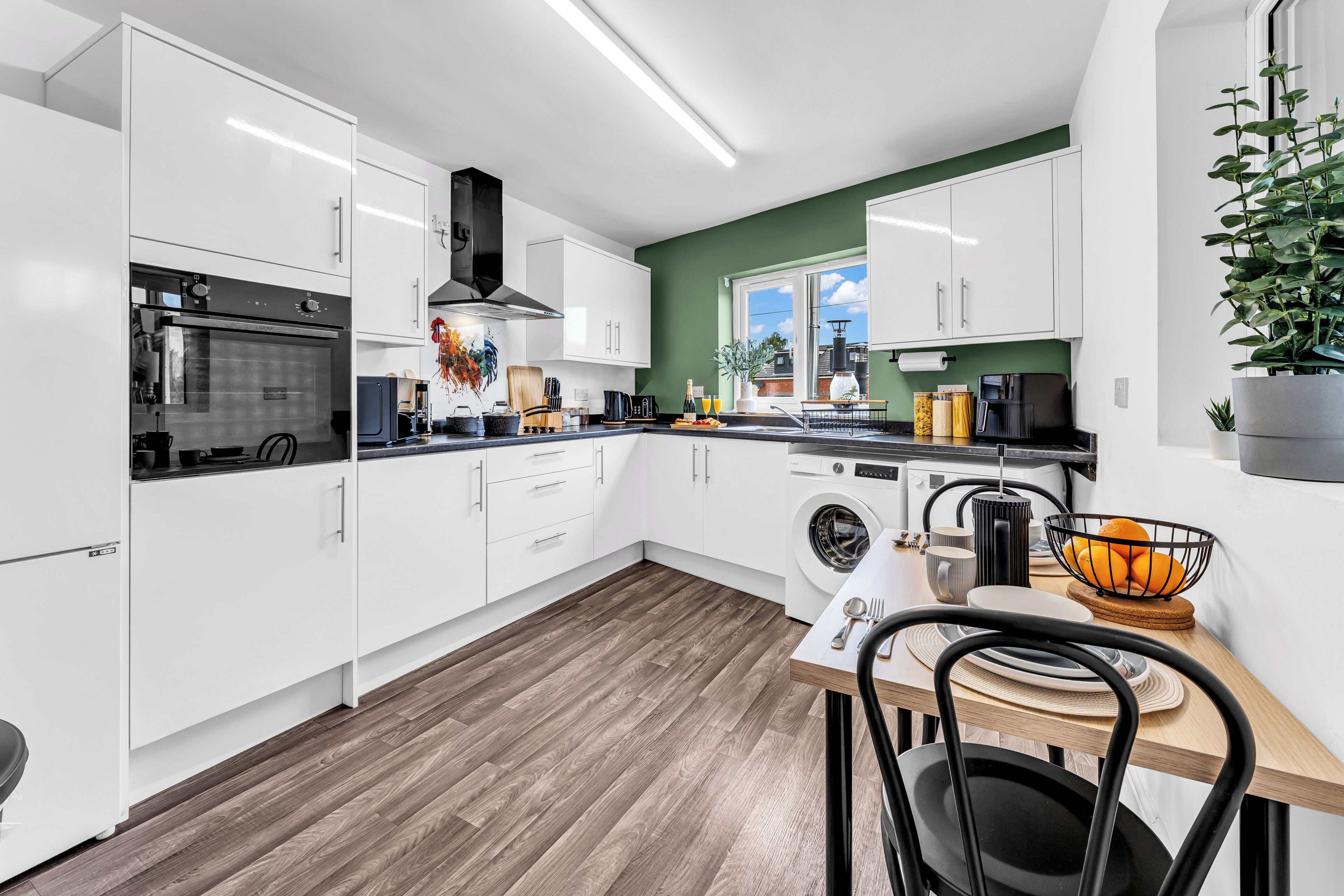 A modern kitchen with white cabinets, a built-in oven, a washing machine, and a dining area with a table and chairs.