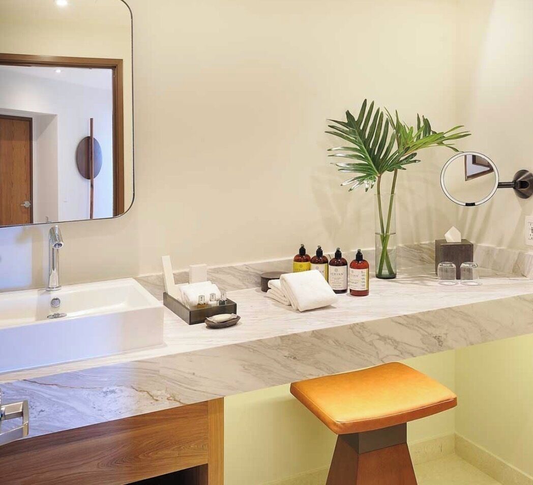 A bathroom with a white sink, a mirror, a round clock, a plant, and toiletries on the counter.