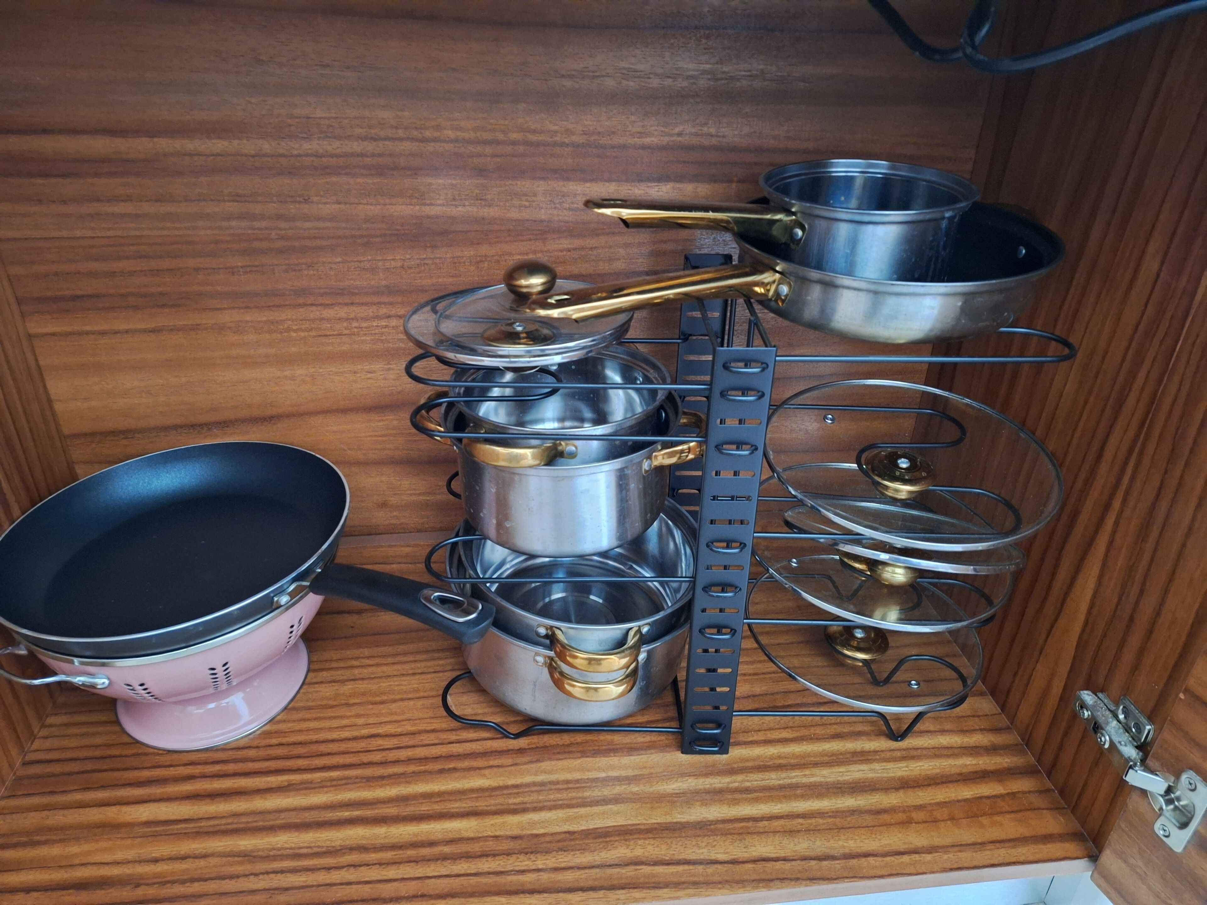 Cozinha com armário de madeira contendo diversas panelas e frigideiras, incluindo um escorredor rosa e uma grade de metal com panelas.