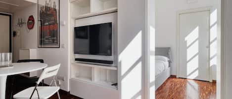 Una habitación moderna con cama, televisor, escritorio blanco con sillas y un cartel en la pared.