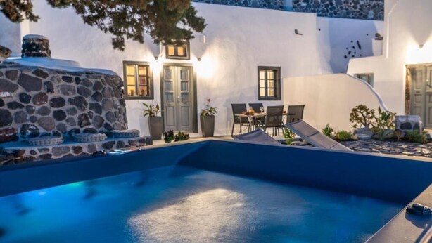 A swimming pool with a stone structure, a dining area with chairs and a table, and a building with a white facade and doors. - Noelia Suites - Cave by ΜΡS (OIA)
