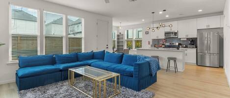 A modern living room with a sectional sofa, a glass coffee table, and a kitchen area with stainless steel appliances.