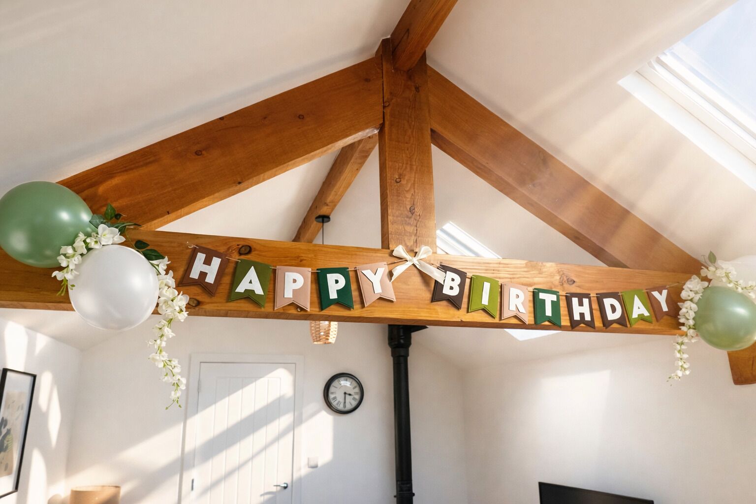 A room with a wooden ceiling, a 'HAPPY BIRTHDAY' banner, balloons, and a clock on the wall.