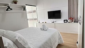 Moderni makuuhuone, jossa on suuri sänky, litteänäyttöinen televisio, pieni pöytä ja ruukussa oleva kasvi.