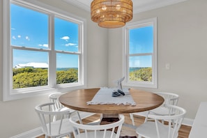 A dining area with a round table, white chairs, and a view through large windows. - New 2025 Build | Private Pool, Elevator | Ocean & Sound Views | DOGS OK (Emerald Isle)