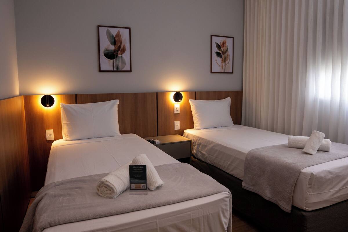 Une chambre d’hôtel avec deux lits, chacun équipé d’un oreiller et d’une serviette enroulée. Des lampes murales et des œuvres d’art encadrées sont disposées au-dessus des lits.