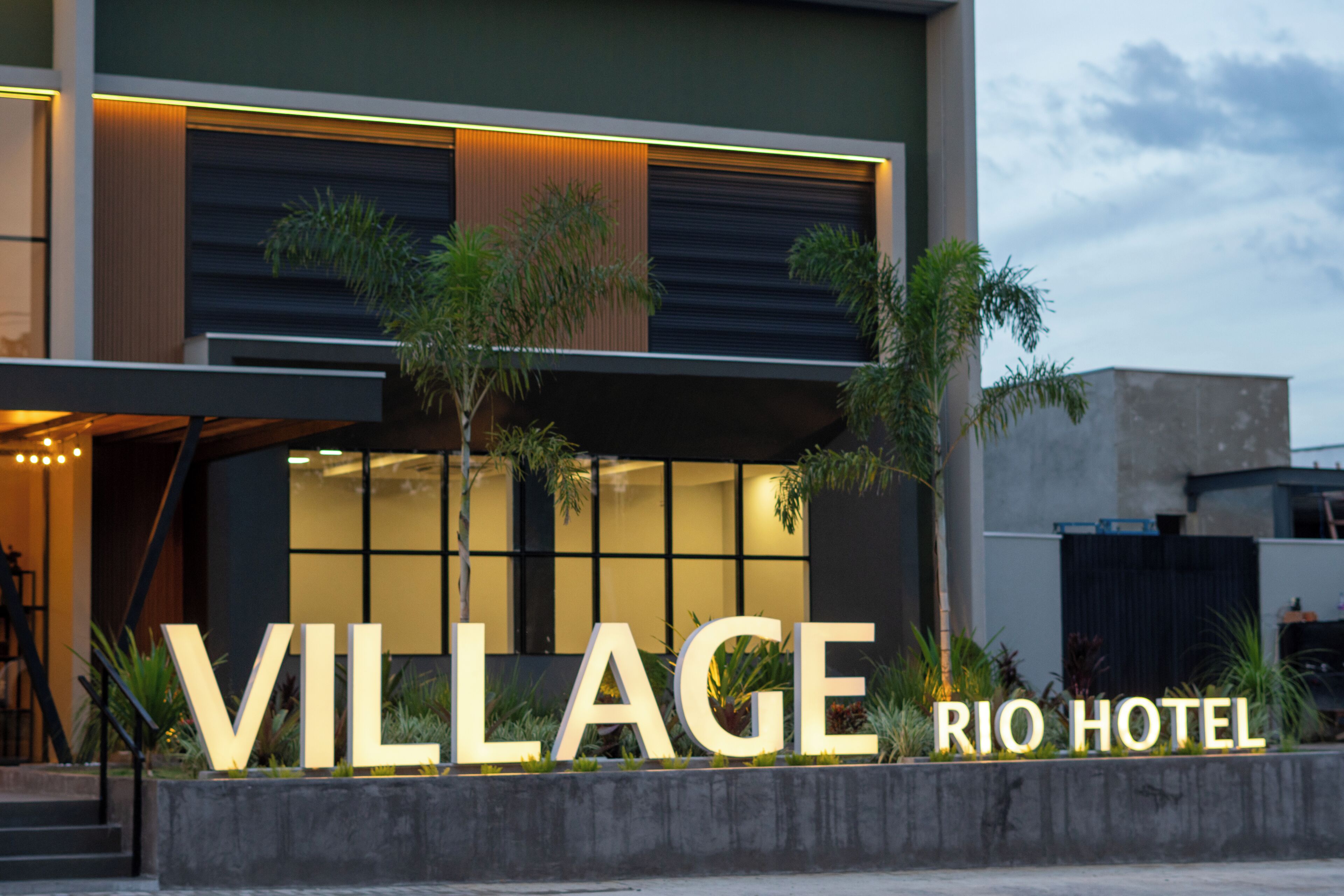 A modern hotel with a prominent 'VILLAGE RIO HOTEL' sign, large windows, and a well-lit entrance.