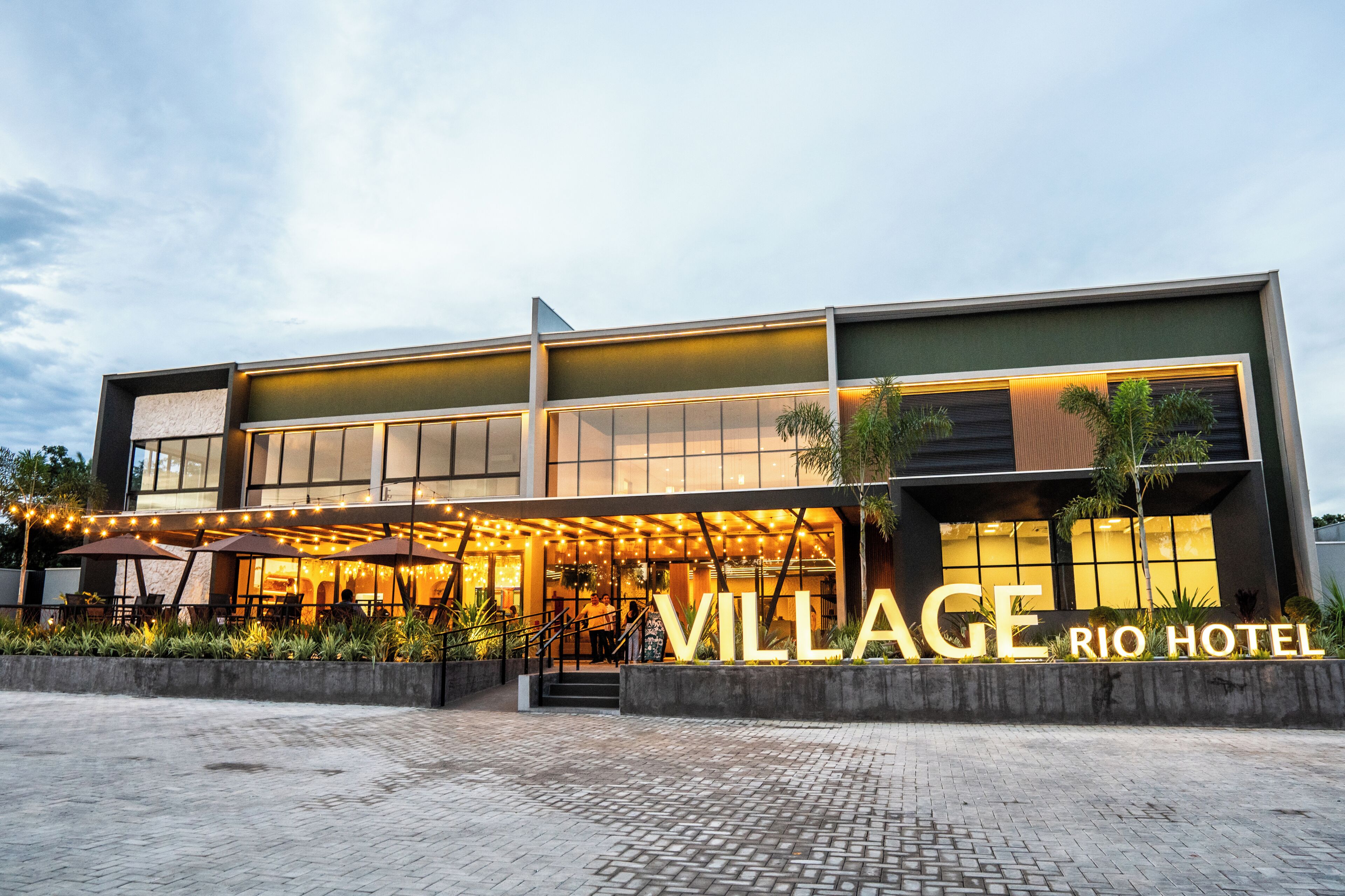 Un hotel moderno con un llamativo letrero que dice 'VILLAGE RIO HOTEL', zona de estar al aire libre y entrada pavimentada.